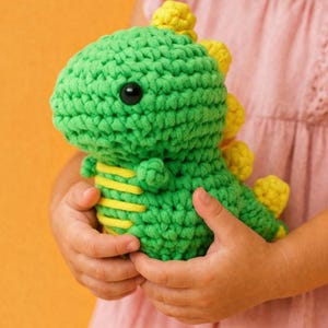以下が含まれることがあります： 手作りの緑色の編み物恐竜のおもちゃ。黄色いアクセントが付いています。恐竜は丸い頭、黒い目、背中に黄色のトゲがあります。黄色の縞模様が体を飾っています。子供の手で持たれています。