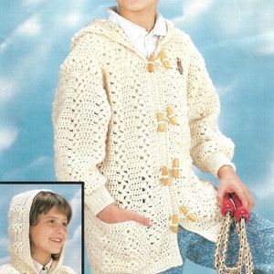 Puede incluir: Un cárdigan blanco de ganchillo con capucha y cierres de botón de madera. El cárdigan tiene un bolsillo en la parte delantera y lo lleva puesto un niño con coletas.