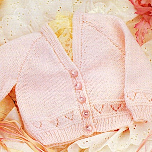 May include: A pink knitted baby cardigan with a V-neck and button fastening. The cardigan features a delicate lace pattern on the sleeves and bottom hem.
