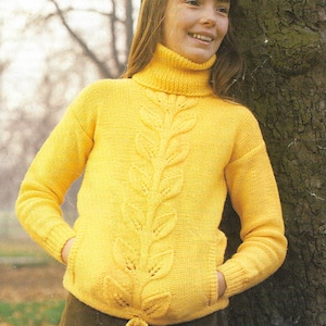 Puede incluir: Un suéter de punto amarillo con cuello alto y un patrón de hojas en la parte delantera. El suéter tiene un lazo en la parte inferior y bolsillos en los lados.