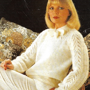 Könnte beinhalten: Ein weißer Strickpullover mit einem zarten Spitzenmuster und einer Schleife am Ausschnitt. Der Pullover wird von einer Frau mit blonden Haaren getragen und sitzt auf einem floral gemusterten Sofa.