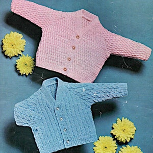May include: Two knitted baby cardigans, one pink and one blue, with button closures. The cardigans are laid out on a blue background with yellow flowers.