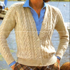 Op de afbeelding: Een crèmekleurige kabelgebreide cardigan met een v-hals en knoopsluiting. De cardigan wordt gedragen over een lichtblauw overhemd met knopen.