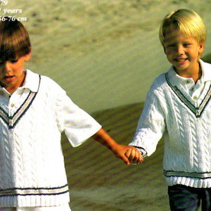 May include: Two young boys wearing white cable knit sweaters with navy blue trim. The sweaters have a v-neck and a button closure. The boys are standing on a sandy beach and holding hands.