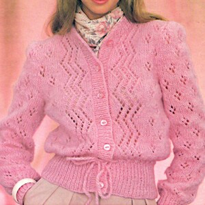 May include: A light pink, open-stitch cardigan with a button-up front and a drawstring waist. The long-sleeved sweater features a textured, open-weave design and a ribbed hem. The cardigan is paired with a floral scarf.
