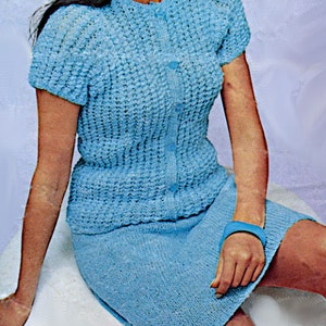 May include: A light blue knitted two-piece outfit. The top is a short-sleeved cardigan with button closures. The skirt is knee-length. The outfit is made of a textured knit fabric. A blue bracelet is visible on the wrist.