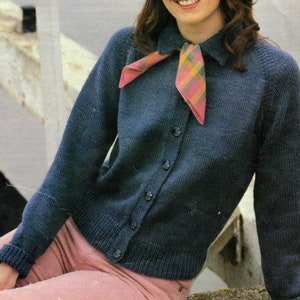 May include: A dark blue knitted cardigan with a button-up front and a collared neckline. The cardigan has long sleeves and a relaxed fit. The model is wearing a pink and yellow plaid scarf tied around the collar.