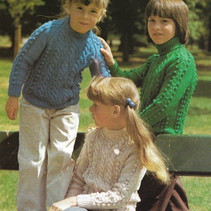 May include: Three children wearing knitted sweaters. The girl on the left is wearing a blue sweater with a cable knit pattern. The girl in the middle is wearing a cream-colored sweater with a cable knit pattern. The girl on the right is wearing a green sweater with a cable knit pattern.