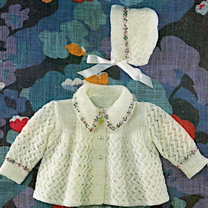 May include: A white knitted baby cardigan with a lace pattern and a matching white knitted hat with a pink and green embroidered trim. The cardigan has a button closure and a collar.