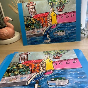 Puede incluir: Dos impresiones art&iacute;sticas que representan un colorido paisaje urbano con edificios, barcos y un cisne sobre agua azul. Los edificios tienen fachadas rojas, naranjas, amarillas y rosas. Las impresiones tienen un borde azul.
