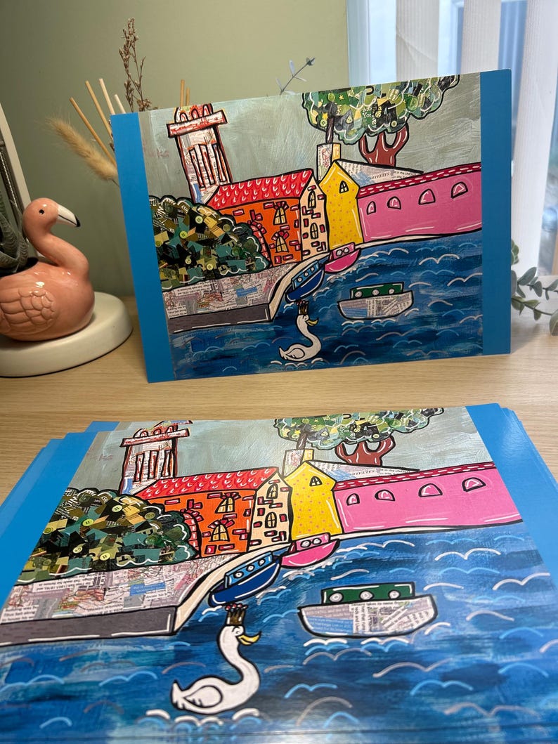 Puede incluir: Dos impresiones art&iacute;sticas que representan un colorido paisaje urbano con edificios, barcos y un cisne sobre agua azul. Los edificios tienen fachadas rojas, naranjas, amarillas y rosas. Las impresiones tienen un borde azul.