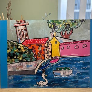 Puede incluir: Obra de arte colorida que representa una escena de un pueblo costero con edificios, barcos y un cisne. Los edificios tienen tejados rojos, naranjas y amarillos. El agua es azul y el cielo es de color gris claro. La obra est&aacute; enmarcada con un borde azul.