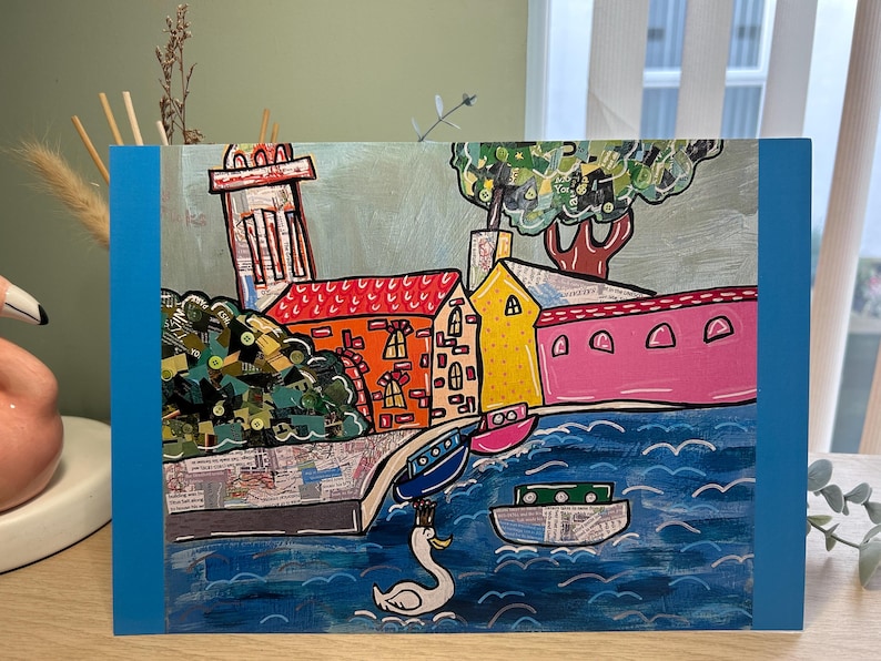 Puede incluir: Obra de arte colorida que representa una escena de un pueblo costero con edificios, barcos y un cisne. Los edificios tienen tejados rojos, naranjas y amarillos. El agua es azul y el cielo es de color gris claro. La obra est&aacute; enmarcada con un borde azul.