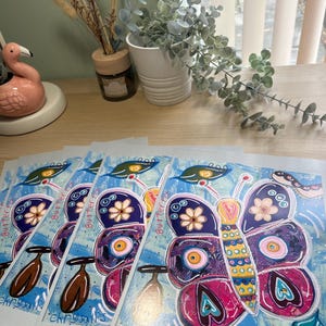 May include: A collection of art prints displaying a vibrant butterfly illustration. The butterfly's wings are a mix of purple, pink, and yellow, adorned with floral and eye motifs. The text "Butterfly Chrysalis" is printed on the artwork.