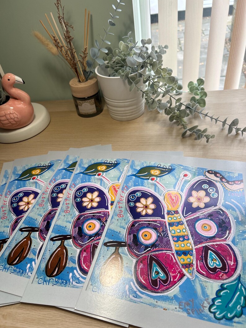 May include: A collection of art prints displaying a vibrant butterfly illustration. The butterfly's wings are a mix of purple, pink, and yellow, adorned with floral and eye motifs. The text "Butterfly Chrysalis" is printed on the artwork.