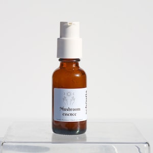 May include: A small amber glass bottle with a white pump dispenser. The label reads "Mushroom essence" with a sun and moon graphic. The bottle sits on a clear, rectangular platform against a white background.