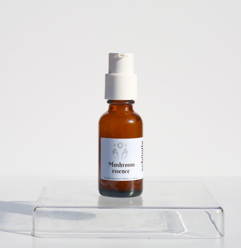 May include: A small amber glass bottle with a white pump dispenser. The label reads "Mushroom essence" with a sun and moon graphic. The bottle sits on a clear, rectangular platform against a white background.