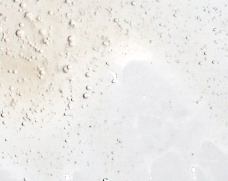 May include: Close-up of a clear, viscous liquid with numerous small bubbles. The liquid has a slightly yellowish tint and a smooth, reflective surface. The bubbles vary in size and are scattered throughout the liquid.