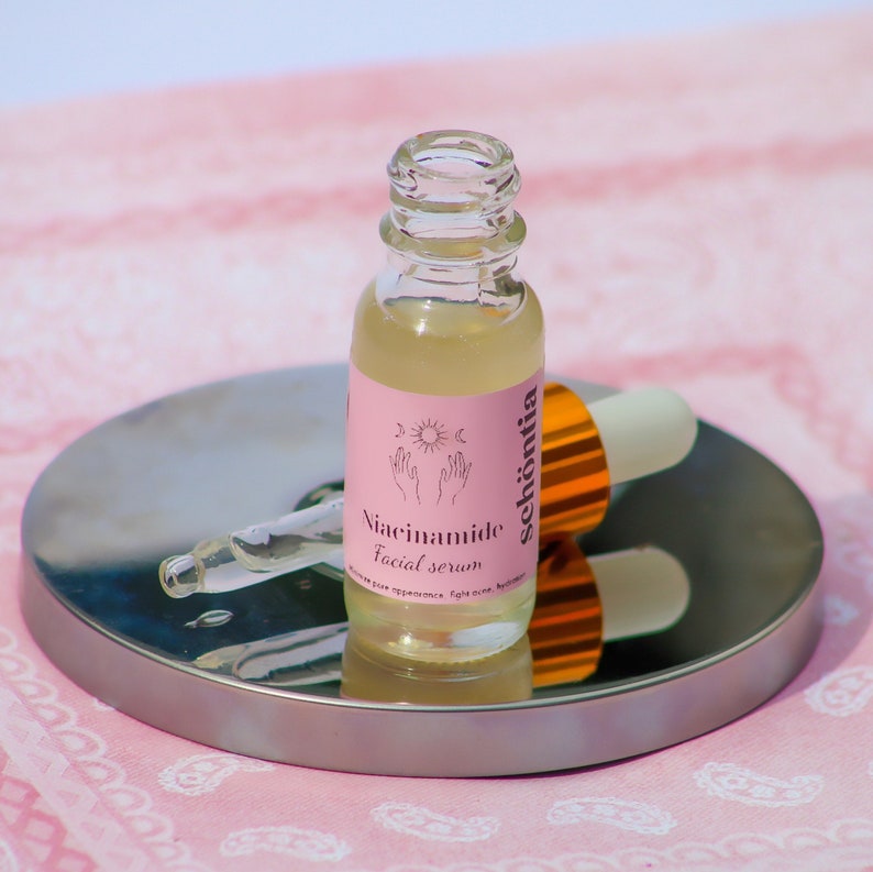 May include: A clear glass bottle of Sch&ouml;nia Niacinamide Facial Serum with a pink label, sun and hand graphic, and a dropper. The bottle sits on a silver tray, with a pink and white patterned background.