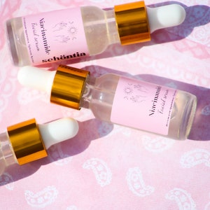 May include: Three clear glass bottles of facial serum with white droppers and gold collars are arranged on a pink paisley fabric. Each bottle has a pink label with the words "Niacinamide Facial Serum" and the brand name "sch&ouml;ntia".