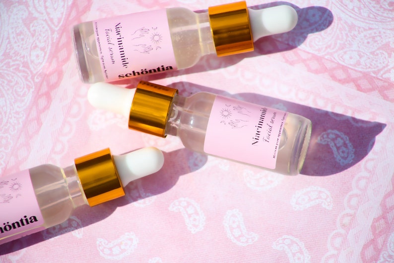 May include: Three clear glass bottles of facial serum with white droppers and gold collars are arranged on a pink paisley fabric. Each bottle has a pink label with the words "Niacinamide Facial Serum" and the brand name "sch&ouml;ntia".