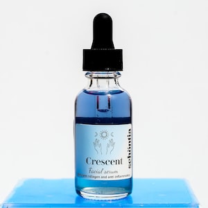 May include: A clear glass bottle with a black dropper filled with blue liquid. The label reads "Crescent Facial serum" with the brand name "sch&ouml;ntia" and a graphic of hands, a crescent moon, and a sun. The bottle sits on a light blue surface.