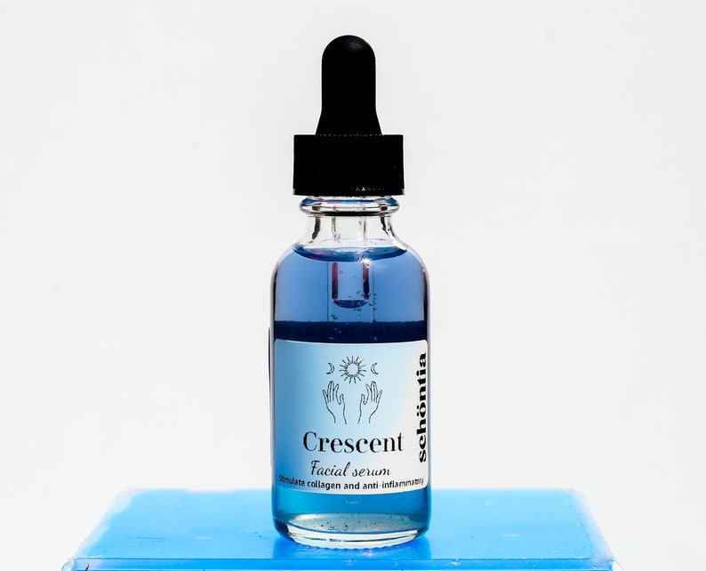 May include: A clear glass bottle with a black dropper filled with blue liquid. The label reads "Crescent Facial serum" with the brand name "sch&ouml;ntia" and a graphic of hands, a crescent moon, and a sun. The bottle sits on a light blue surface.