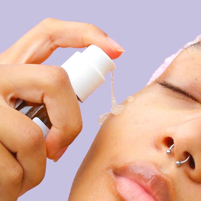 May include: A person applying a clear gel from a white pump bottle to their face. The gel is dispensed onto the skin, with a light purple background. The bottle has a brown label.