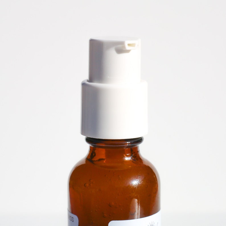 May include: Close-up of a brown glass bottle with a white pump dispenser. The bottle has a rounded shape and a short neck. The pump is white with a small spout. The background is plain white.