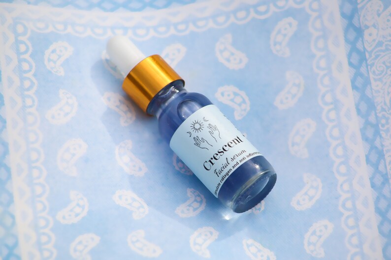 May include: A small glass bottle of Crescent Facial Serum with a blue liquid inside. The bottle has a white dropper top with a gold band. The label is light blue with the words "Crescent Facial Serum" and a small illustration.
