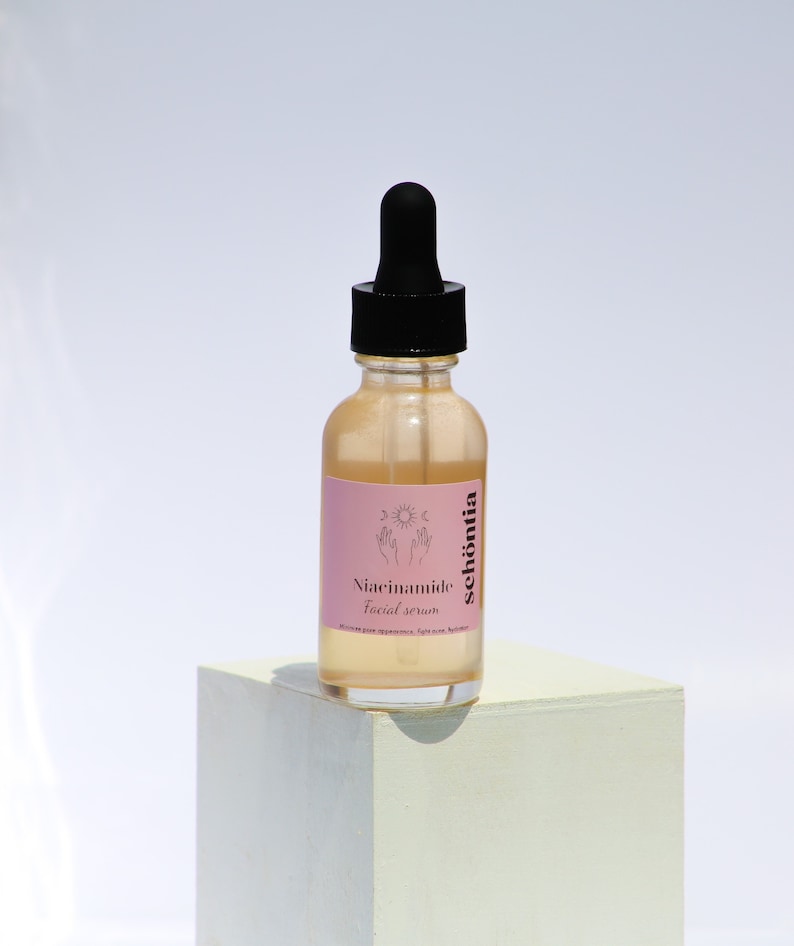 May include: A clear glass bottle with a black dropper top, containing a light-colored serum. The bottle has a pink label with the text "Niacinamide Facial Serum" and the brand name "sch&ouml;ntia". The bottle is placed on a white cube.
