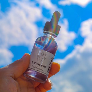 May include: A clear glass bottle of facial serum, held against a bright blue sky with fluffy white clouds. The bottle has a dropper and a white label with the text "Crescent Facial serum" and the brand name "sch&ouml;ntia". The serum appears to be a light purple color.