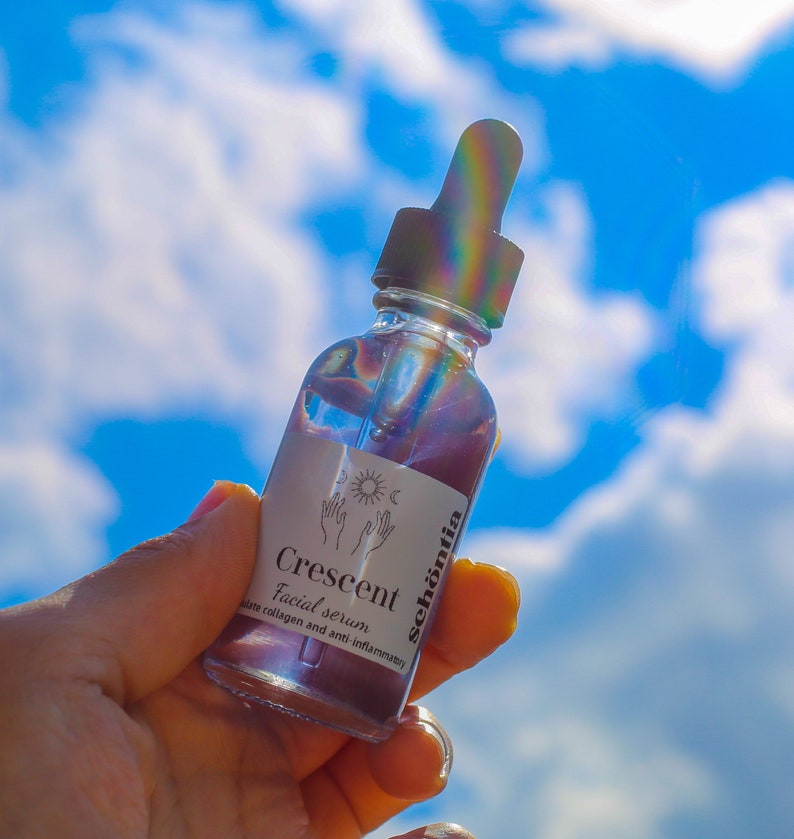 May include: A clear glass bottle of facial serum, held against a bright blue sky with fluffy white clouds. The bottle has a dropper and a white label with the text "Crescent Facial serum" and the brand name "sch&ouml;ntia". The serum appears to be a light purple color.