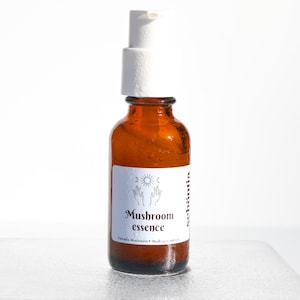May include: Amber glass bottle with a white pump dispenser. The label reads "Mushroom essence" with the words "Tremella Mushroom + Mushroom extract" below. The label also features a sun, moon, and hand design.
