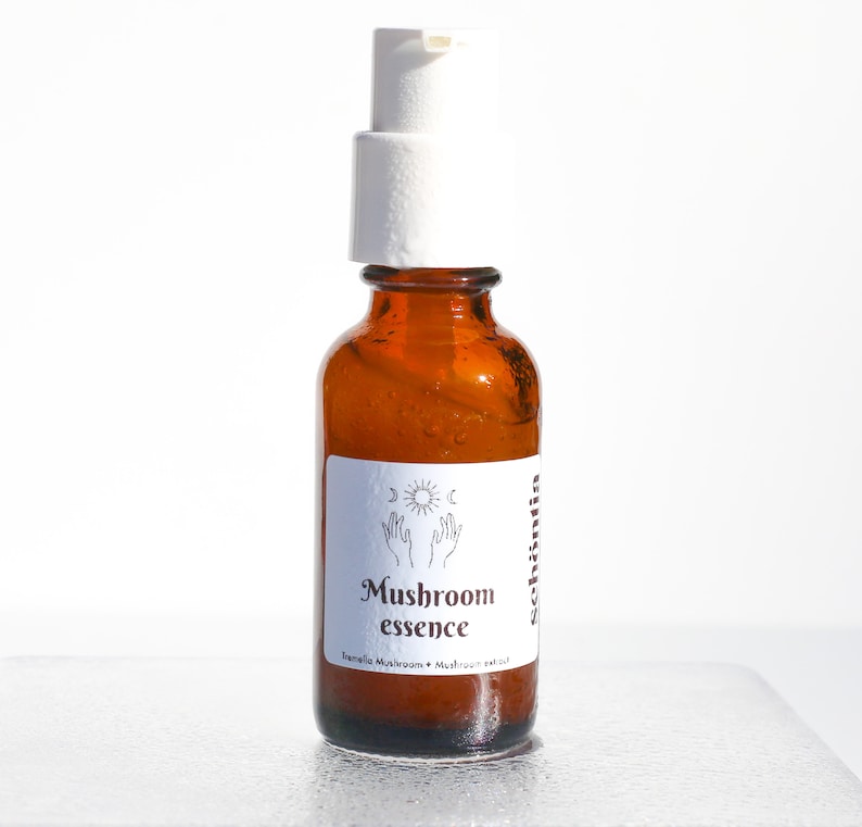 May include: Amber glass bottle with a white pump dispenser. The label reads "Mushroom essence" with the words "Tremella Mushroom + Mushroom extract" below. The label also features a sun, moon, and hand design.