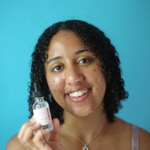 May include: A clear glass bottle with a dropper, labeled "sebonita" in pink text. The bottle contains a clear liquid. The background is a solid turquoise color. The product is held in front of a person.