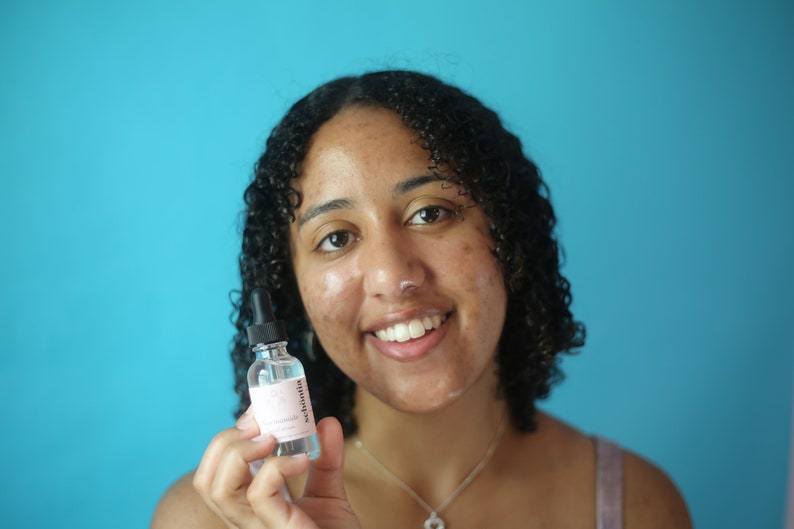 May include: A clear glass bottle with a dropper, labeled "sebonita" in pink text. The bottle contains a clear liquid. The background is a solid turquoise color. The product is held in front of a person.