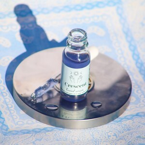 May include: A small glass bottle of "Crescent" facial serum with a dropper, filled with a purple liquid. The bottle has a white label with a sun, moon, and hand design. The bottle sits on a silver metal surface.