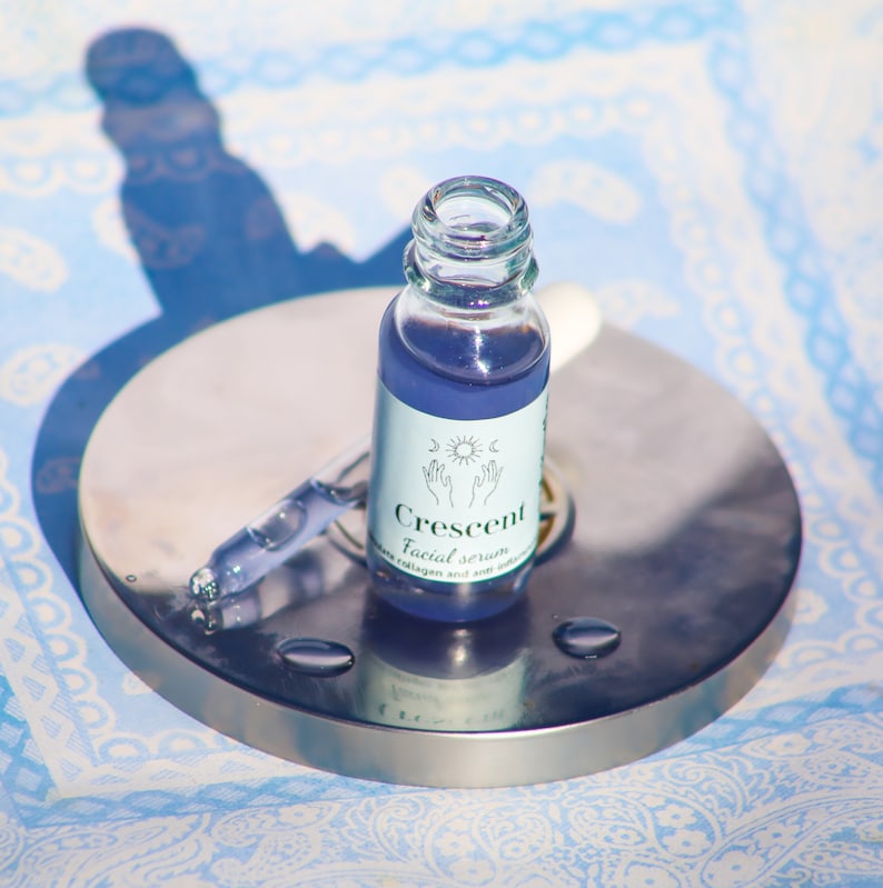 May include: A small glass bottle of "Crescent" facial serum with a dropper, filled with a purple liquid. The bottle has a white label with a sun, moon, and hand design. The bottle sits on a silver metal surface.
