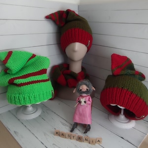 May include: Hand-knitted elf hats and a matching scarf in green, red, and olive green. The hats have a pointed top and a red stripe. A small doll in a pink dress is in front of the hats. The word "KASTANIA" is spelled out in wooden letter tiles.