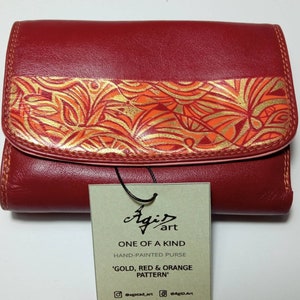 May include: A red leather wallet with a gold, red and orange hand-painted design. The wallet has a flap closure and a tag that reads "Agi art, One of a Kind, Hand-Painted Purse, 'Gold, Red & Orange Pattern', @agatad.art, @AgID.Art".