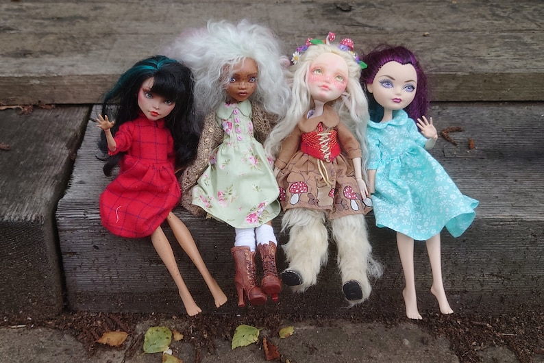 May include: Four dolls sitting on wooden steps. The dolls are wearing different dresses and have different hair colors and styles. The doll on the left is wearing a red dress with a plaid pattern. The doll in the middle is wearing a green dress with a floral pattern. The doll on the right is wearing a blue dress with a floral pattern. The doll in the back is wearing a white dress with a floral pattern.