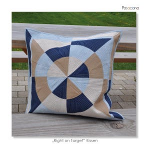 Könnte beinhalten: Ein quadratisches Zierkissen mit geometrischem Design. Das Kissen hat ein kreisförmiges Muster in Blau-, Beige- und Cremetönen mit gestickten Linien. Der Text "Right on Target" Kissen befindet sich unten.