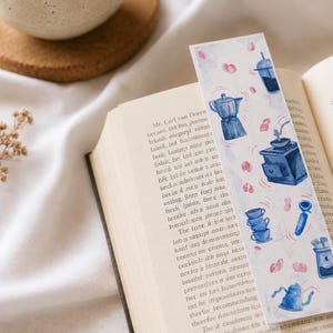 Op de afbeelding: Een rechthoekige papieren boekenlegger met een aquarelillustratie van diverse blauwe koffiezetapparatuur, waaronder een mokkapot, een koffiemolen, gestapelde kopjes en een filterdrager, geaccentueerd met kleine roze cirkelvormige figuren.