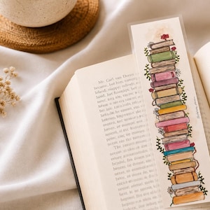 Pode incluir: Um marcador de página retangular com uma pilha vertical de livros ilustrados em várias cores, incluindo verde, rosa, amarelo e azul, acentuado com pequenas vinhas verdes e flores minúsculas, colocado dentro de um livro aberto com texto.