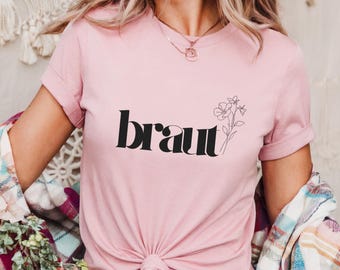 Puede incluir: Una camiseta rosa con la palabra "braut" en texto negro y un dibujo simple de una flor.
