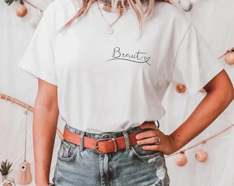 May include: White t-shirt with the word "Braut" written in cursive with a small heart next to it. The shirt is being worn by a person with a brown belt and blue jeans.