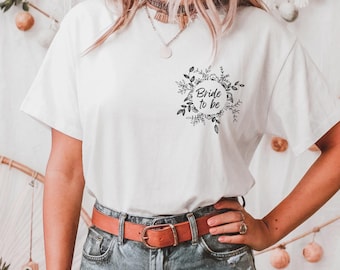 May include: White t-shirt with a black floral wreath design and the text "Bride to be". The shirt has short sleeves and is paired with denim shorts and a brown belt.
