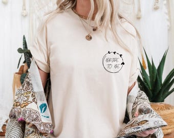 Puede incluir: Una camiseta blanca con el texto "Bride to Be" en un círculo rodeado de corazones.