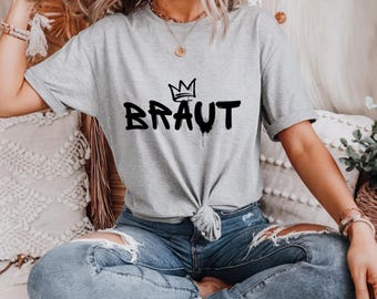 Puede incluir: Una camiseta gris con una corona negra y la palabra "BRAUT" escrita en estilo graffiti.